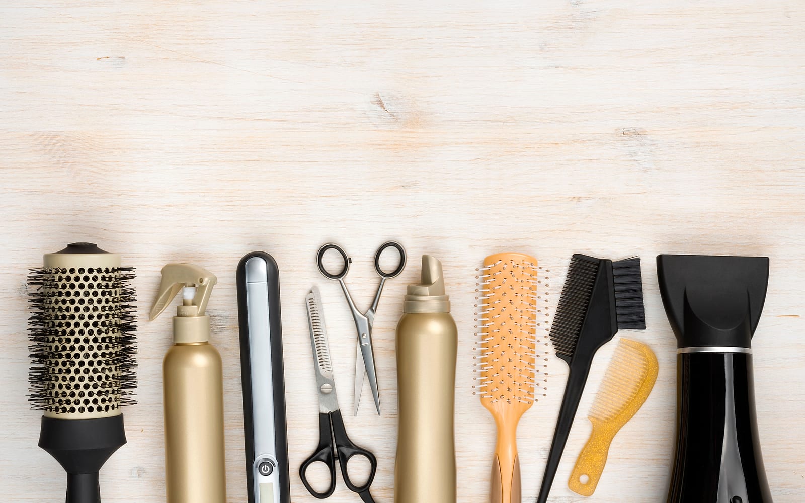 Brushes, scissors, a flat iron, styling sprays and a blow dryer laid out in a row on pale wood